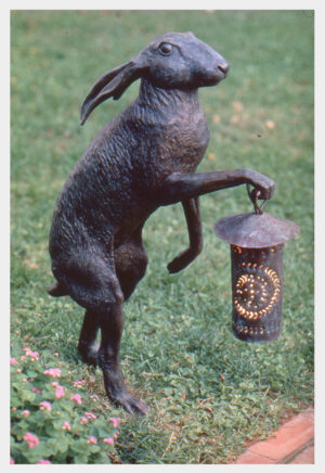 Realistic bronze sculpture of a jack rabbit standing on its hind legs holding an old-fashioned punched copper lantern which has been electrified in its right front paw